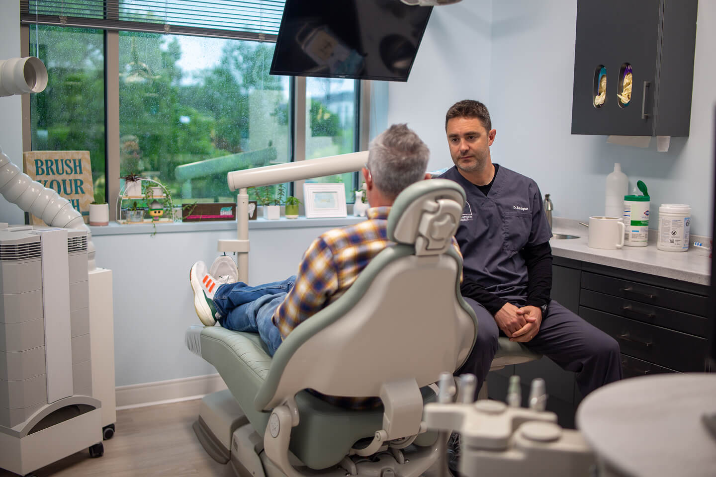 A dentist consults with a patient in a dental office. The patient, in a plaid shirt, sits in a dental chair. The room is calm with equipment visible.